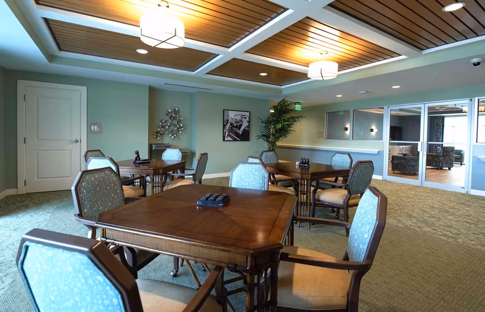 A well-lit common area with several wooden tables and cushioned chairs arranged for group seating. The room features a green carpet, light green walls, decorative ceiling lights, a potted plant, and a framed black and white photograph on the wall. Glass doors lead to another room with additional seating.