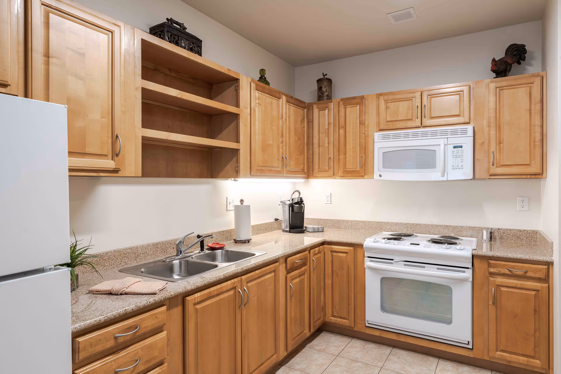 A kitchen with light wood cabinets, a white refrigerator, a white microwave mounted above a white electric stove, a double sink with a faucet, and a beige countertop. There are some decorative items on top of the cabinets and a small plant on the counter near the refrigerator.
