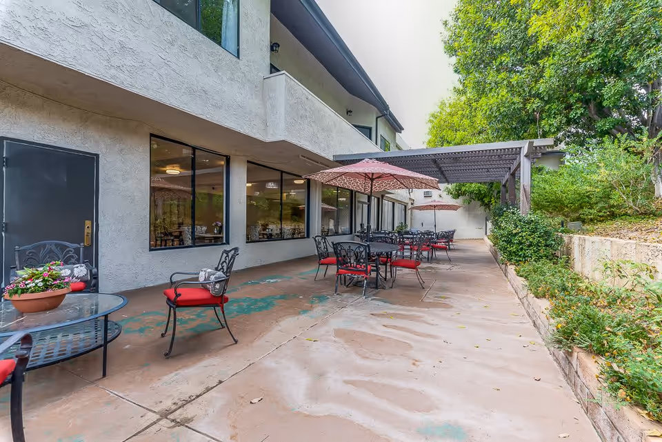 Outdoor patio area at Northridge Valley Senior Living with several black metal tables and chairs with red cushions. Some tables have red umbrellas providing shade. The patio is adjacent to a building with large windows and surrounded by greenery and trees.