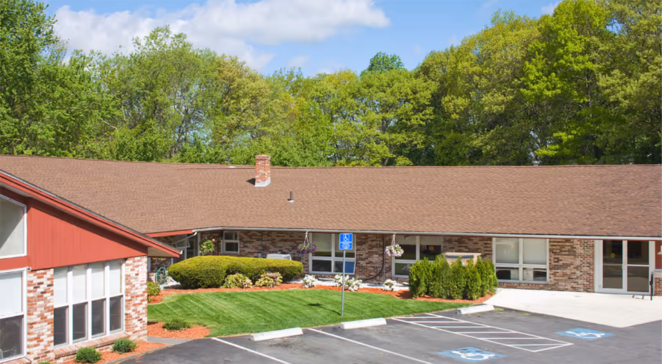 Exterior view of a single-story senior living facility building with a brown shingle roof, brick walls, and large windows. The foreground shows a parking lot with designated handicapped parking spaces and a well-maintained lawn with bushes and hanging flower baskets. Trees and a blue sky with some clouds are visible in the background.