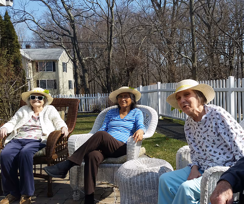Three elderly women sitting outdoors on wicker chairs in a garden area with a white picket fence and leafless trees in the background. They are wearing sun hats and casual clothing, enjoying a sunny day.
