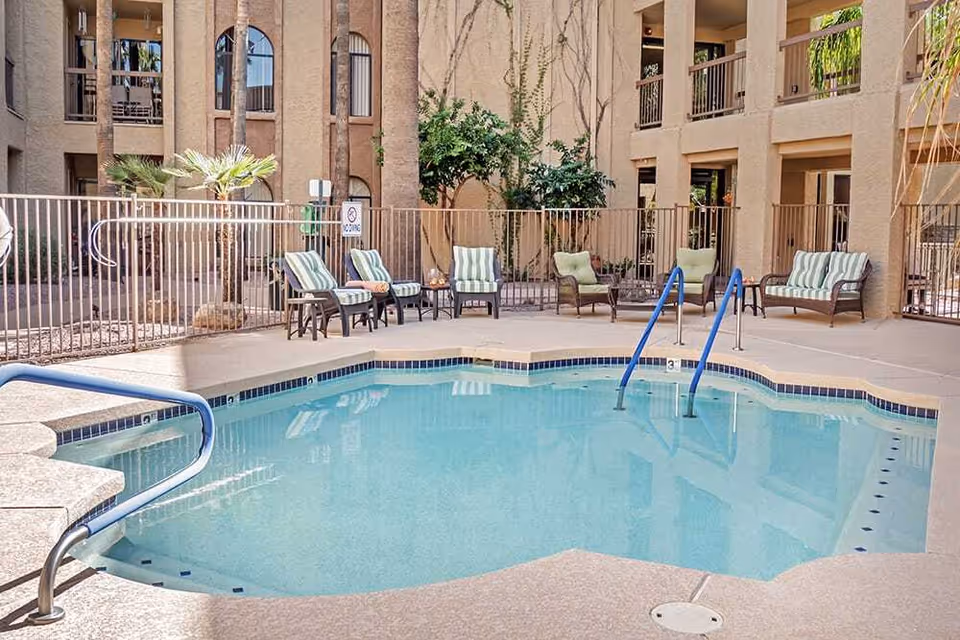 Outdoor swimming pool area at a senior living facility with several cushioned lounge chairs and small tables arranged around the pool. The pool has metal handrails and is surrounded by a safety fence. The building exterior is visible in the background with balconies and palm trees.