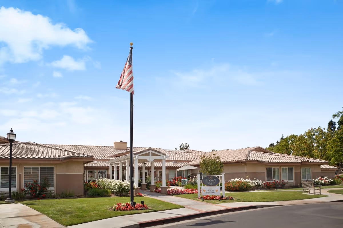 Exterior front view of a single-story Sunrise at Canyon Crest senior living building with a flagpole, landscaped lawns, and an entrance sign.