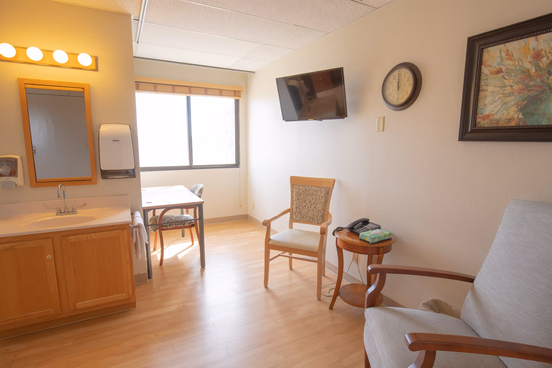 Well-lit senior living room featuring a sink and vanity, a small table by a window, a wall-mounted TV, and two chairs.