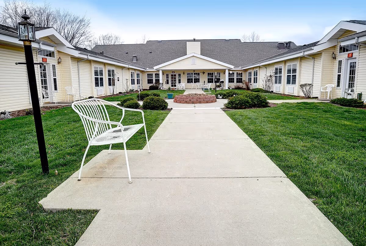 Outdoor courtyard area of a senior living facility with a concrete walkway, green grass, bushes, white metal benches, and a central fire pit. The building surrounds the courtyard with multiple windows and doors leading outside.