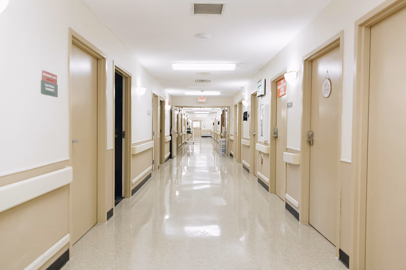 A long, clean hallway in a healthcare facility with beige doors on both sides, handrails along the walls, and bright overhead lighting.