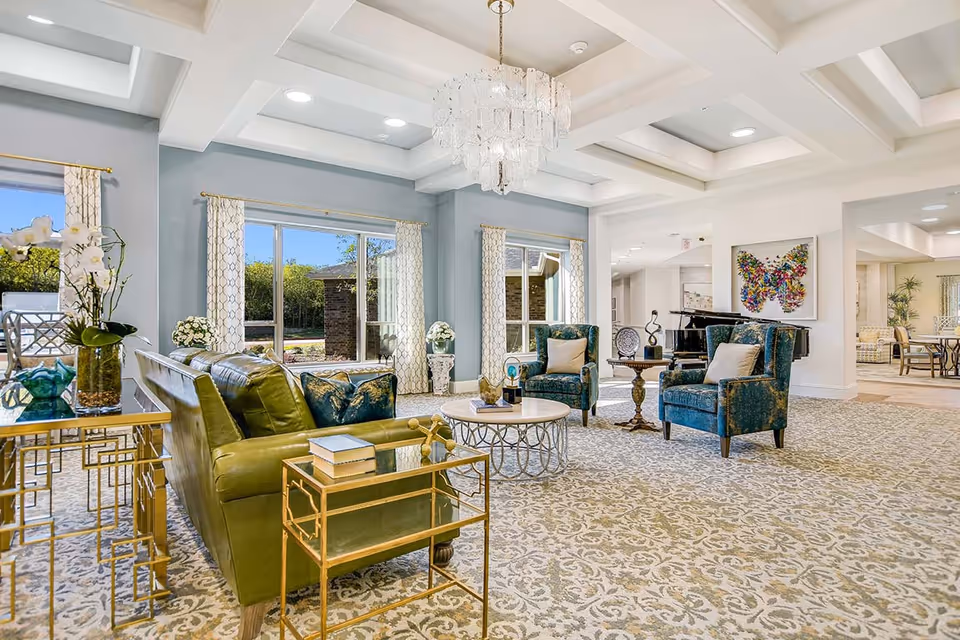 A spacious and elegant living room area with a green leather sofa, two blue upholstered armchairs, a round coffee table, and a side table with books. The room has large windows with patterned curtains letting in natural light, a chandelier hanging from a coffered ceiling, and decorative artwork including a colorful butterfly on the wall. The carpet has a detailed floral pattern, and there is a piano in the background.