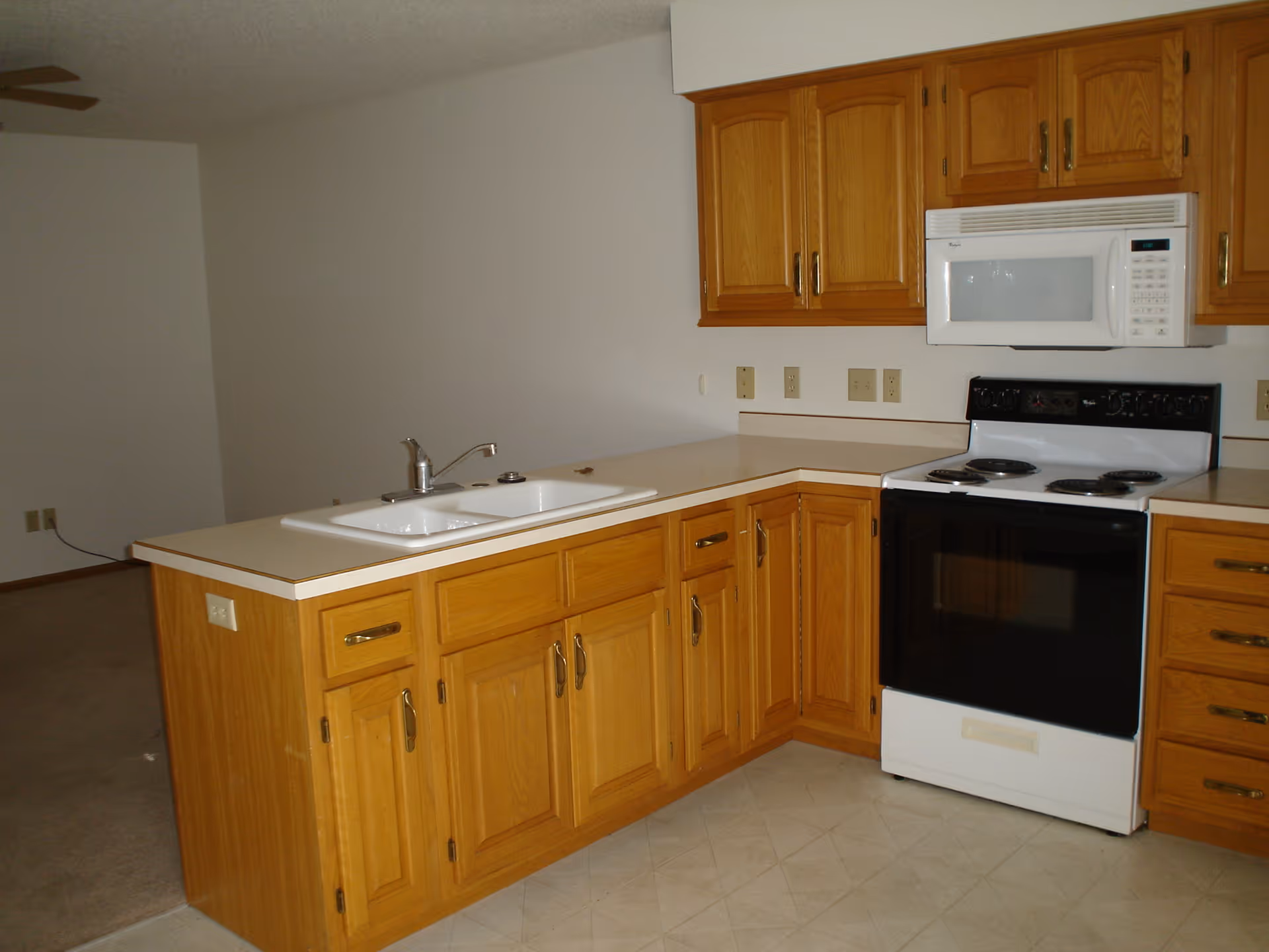 Interior view of a kitchen with wooden cabinets, a white countertop with a double sink, an electric stove with four burners, and a white microwave mounted above the stove. The kitchen opens into a carpeted living area with a ceiling fan.