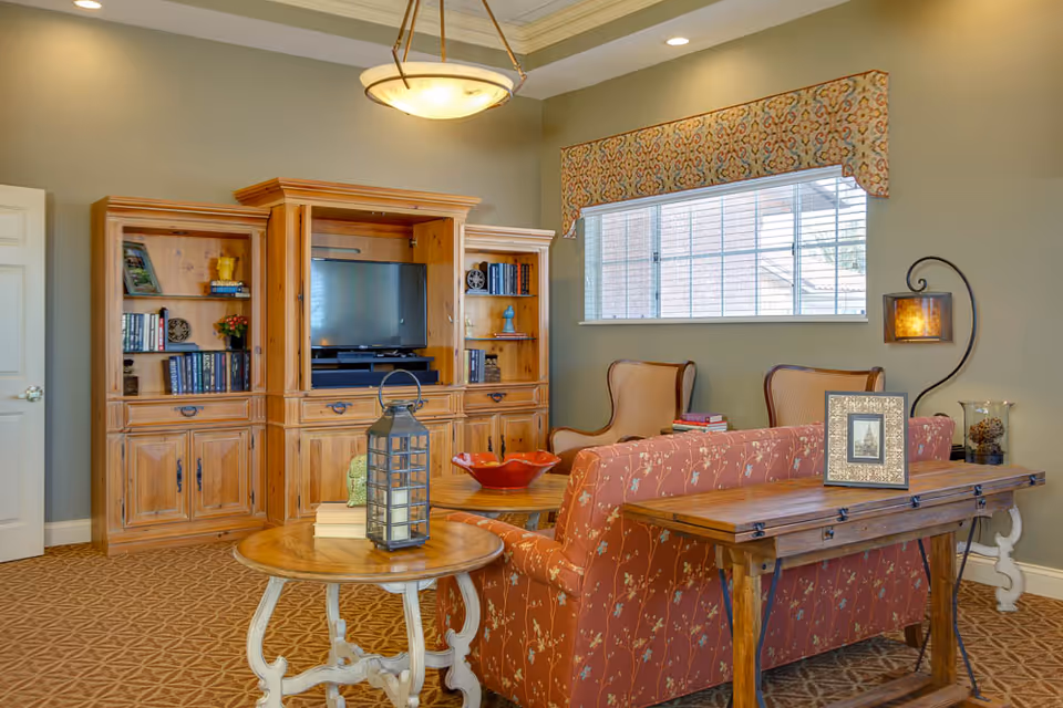 Comfortable living room with a patterned sofa, wooden entertainment center with TV, armchairs, and decorative tables under a pendant light.
