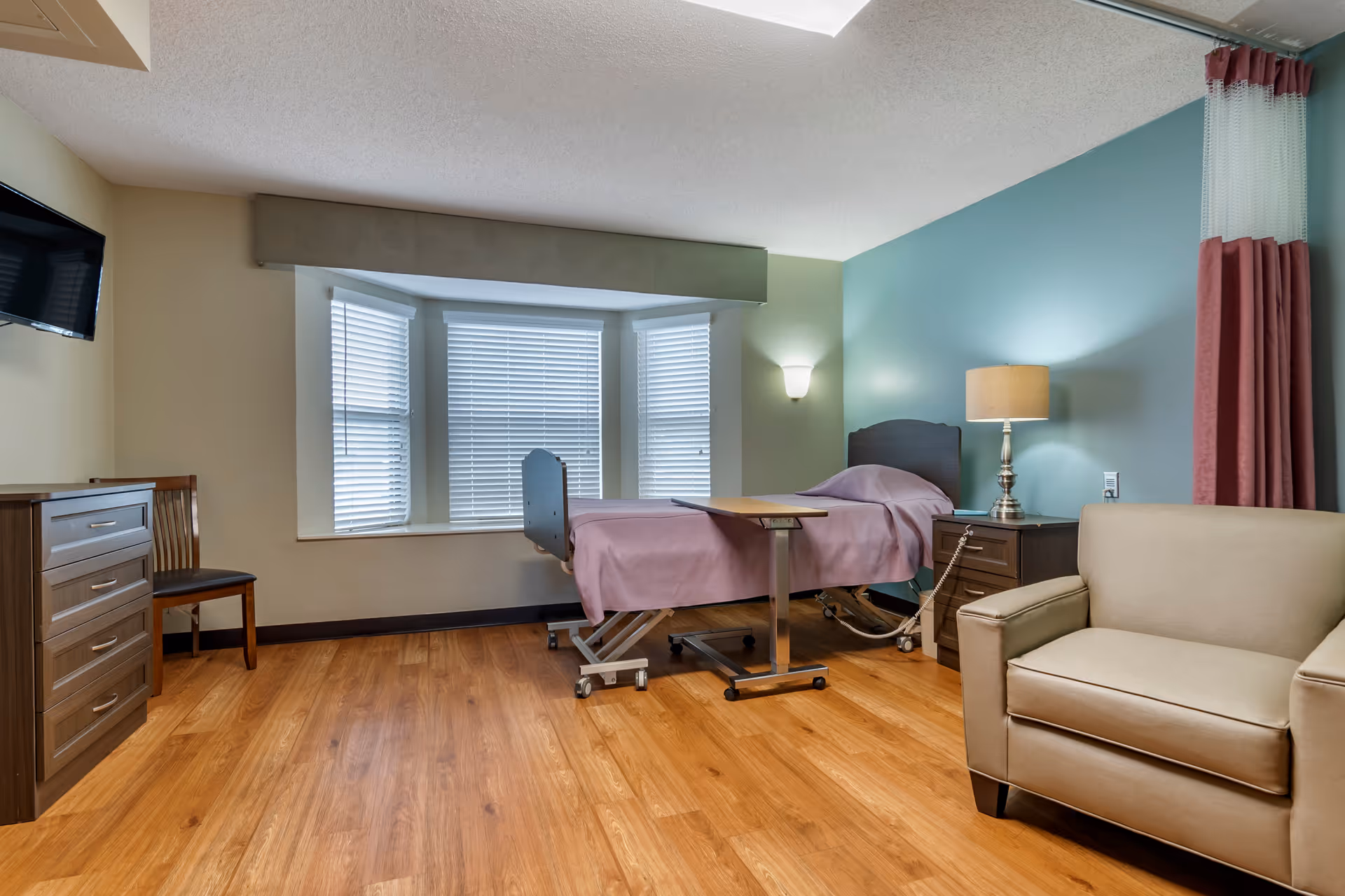 A furnished patient bedroom with a hospital bed, overbed table, armchair, dresser, and wall-mounted TV.
