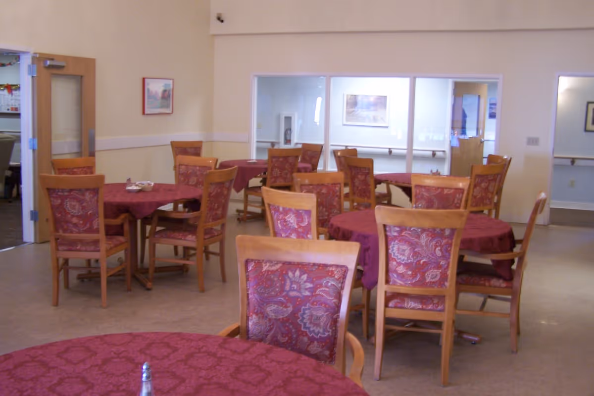 Interior view of a dining room with multiple round tables covered with red patterned tablecloths and wooden chairs with matching red patterned cushions. The room has beige walls, a door on the left, and windows on the far wall.