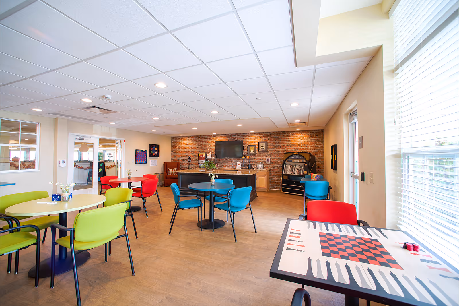 A bright and spacious common area with colorful chairs and round tables, a brick accent wall with a mounted TV, a counter with a coffee machine, and a game table with a checkerboard pattern near a window with blinds.