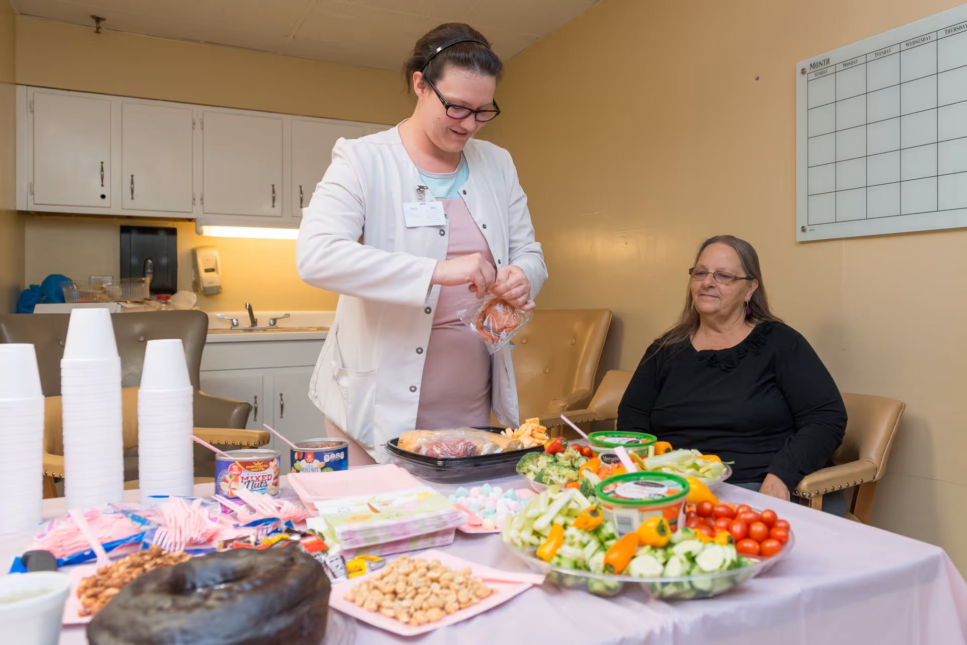 A woman in a white coat is preparing food at a table covered with a pink tablecloth, which has various snacks, vegetables, and cups on it. Another woman with glasses and long hair is sitting on a chair nearby, watching. The setting appears to be a room with beige walls and cabinets in the background.