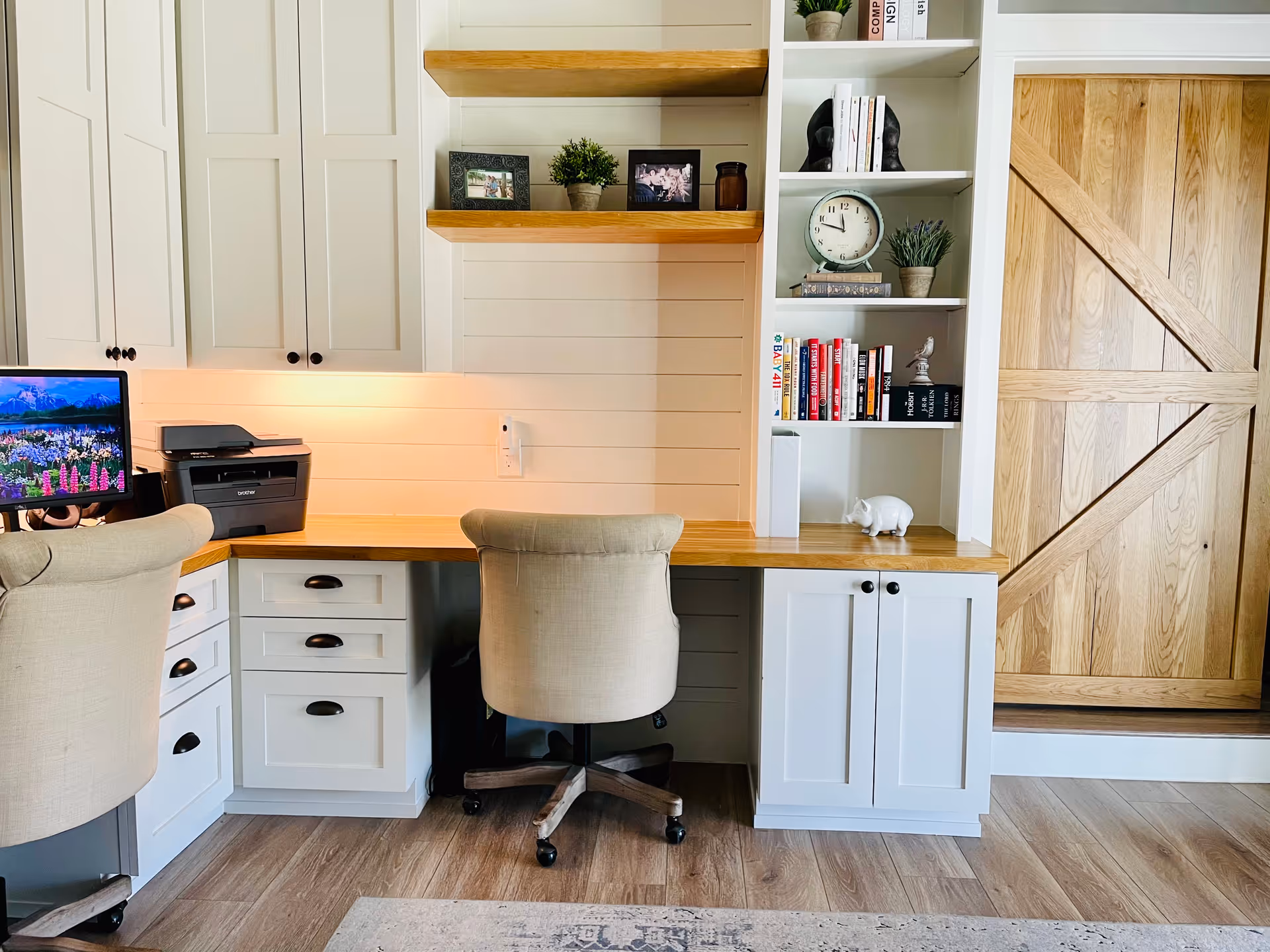 A bright and organized home office space with white cabinetry and wooden countertops. Two beige upholstered office chairs are positioned at the desks. The left desk has a computer monitor displaying a scenic mountain and flower image and a printer. The right side features built-in shelves with books, framed photos, plants, a clock, and decorative items. A wooden barn-style sliding door is visible on the right side of the image. The floor is light wood, and the walls are white with horizontal paneling.