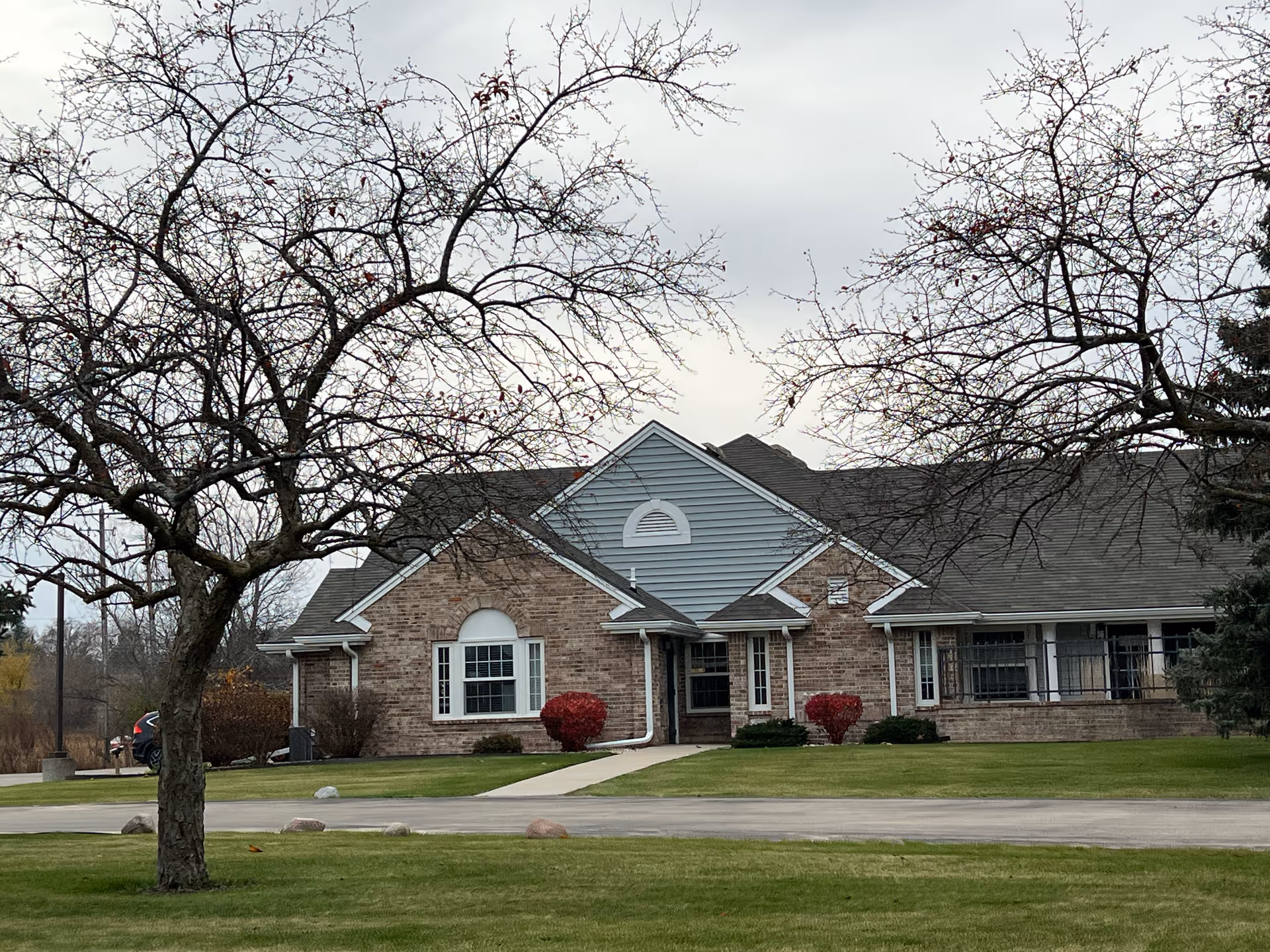 Exterior view of a single-story brick building with a gray roof and white-trimmed windows, surrounded by a well-maintained lawn and leafless trees under a cloudy sky.