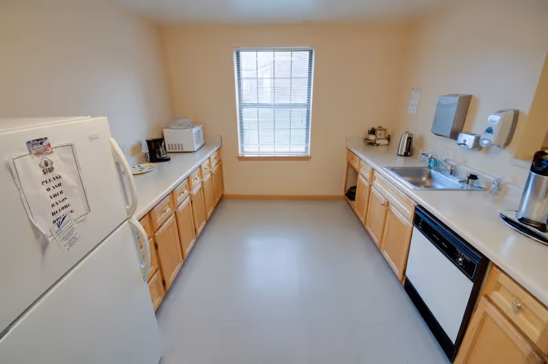 A clean, well-lit kitchen with light wood cabinets and white countertops on both sides. On the left side, there is a white refrigerator with a notice attached, a coffee maker, and a microwave. On the right side, there is a stainless steel sink, a dishwasher, a kettle, and a soap dispenser mounted on the wall. A window with blinds is centered on the far wall, allowing natural light into the room.