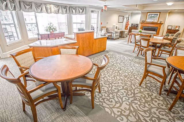 A bright and spacious common area in a senior living facility featuring multiple round wooden tables with chairs arranged around them. The room has large windows with blinds and valances, a patterned carpet, and a wooden reception desk or counter. In the background, there is a seating area with armchairs, a fireplace, framed artwork, and an American flag.