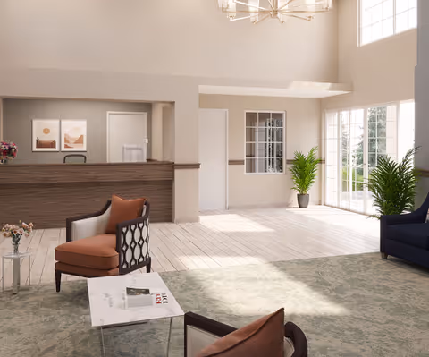 A bright and spacious senior living facility lobby with a wooden reception desk, two framed artworks on the wall behind it, a seating area with cushioned chairs and a marble-top table, large windows allowing natural light, and potted plants near the entrance.