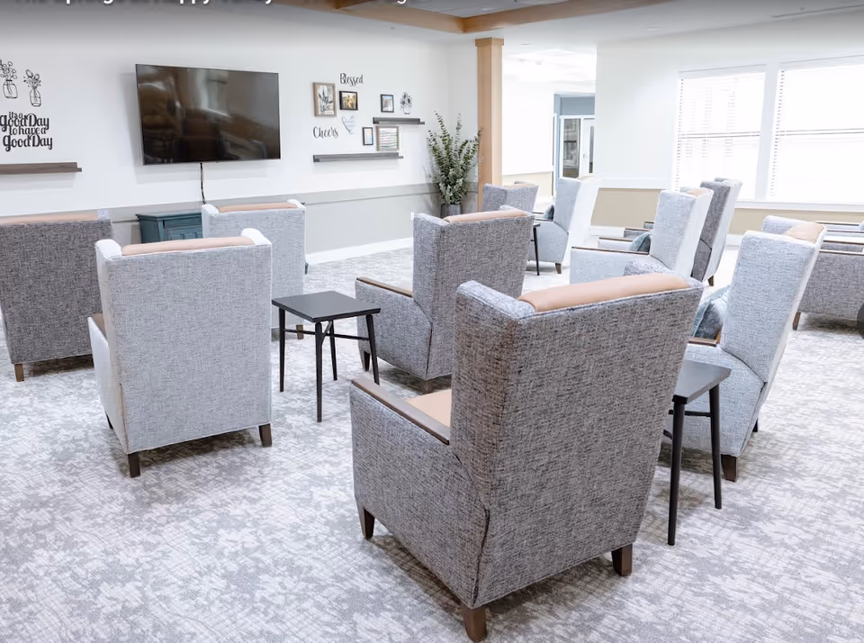 A bright and spacious common area with multiple gray upholstered armchairs arranged in rows facing a wall-mounted flat-screen TV. The room has light-colored walls with decorative shelves and framed pictures, a patterned carpet, and large windows letting in natural light.