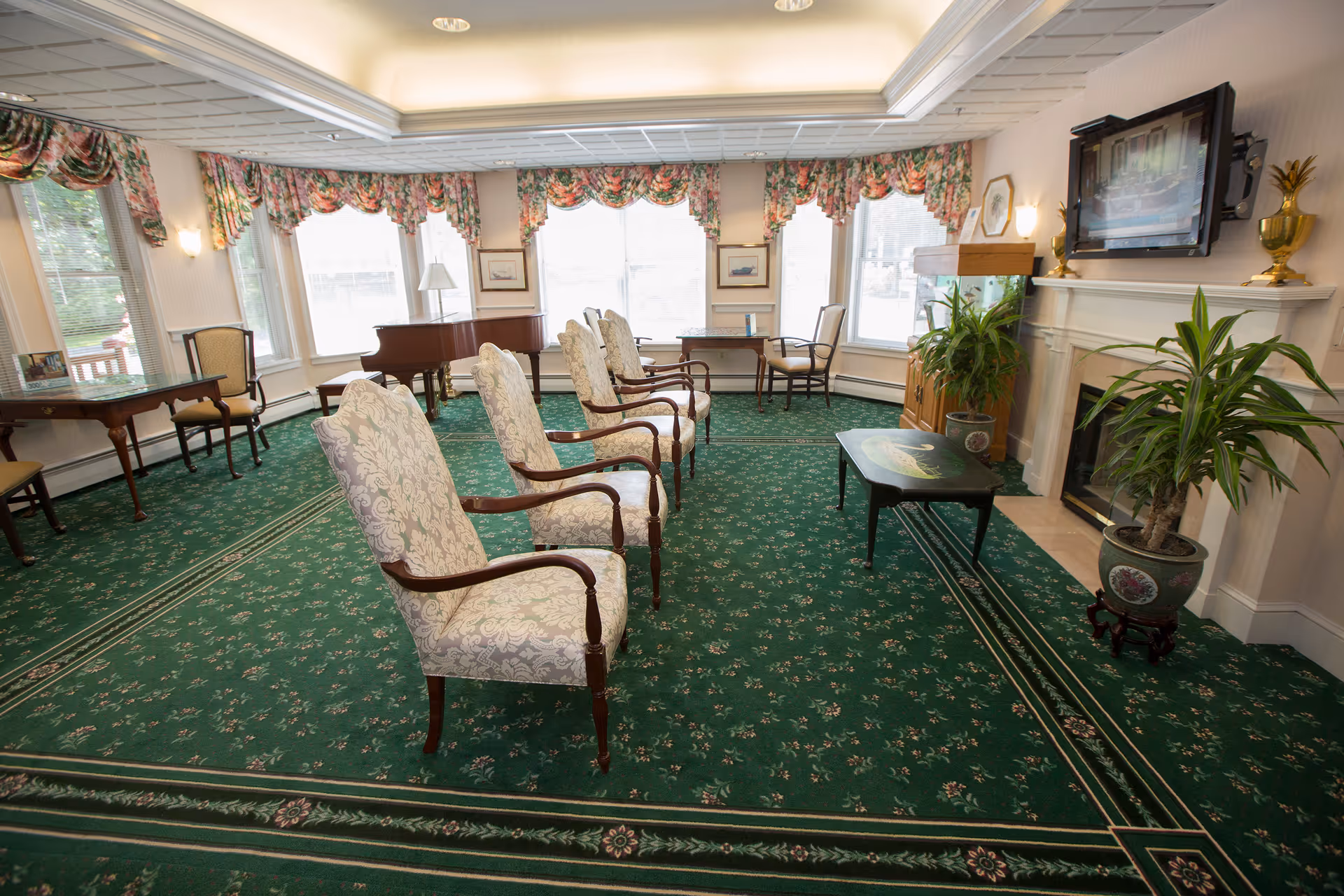 Bright common sitting room with a row of upholstered armchairs, a fireplace and wall-mounted TV, a piano near bay windows, and potted plants.