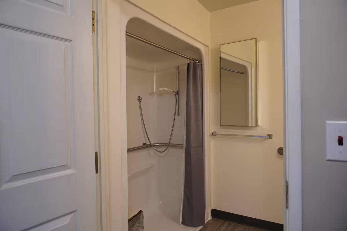 A small bathroom featuring a white shower stall with a gray shower curtain, a handheld showerhead, a wall-mounted mirror above a towel rack, and a light switch on the wall. The door to the bathroom is partially open.