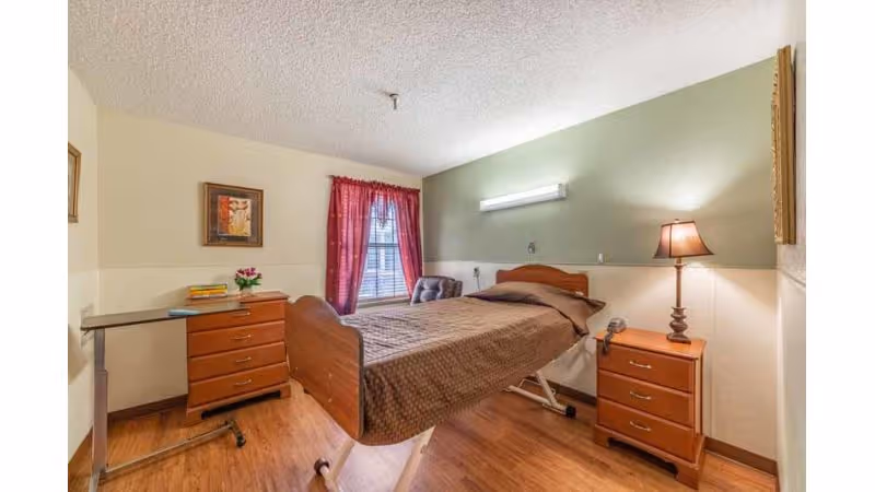 A nursing facility room with a single hospital-style bed covered with a brown blanket. The room has wooden flooring, a green accent wall behind the bed, a bedside table with a lamp, a chest of drawers with flowers and books, a window with red curtains, and a small overbed table.