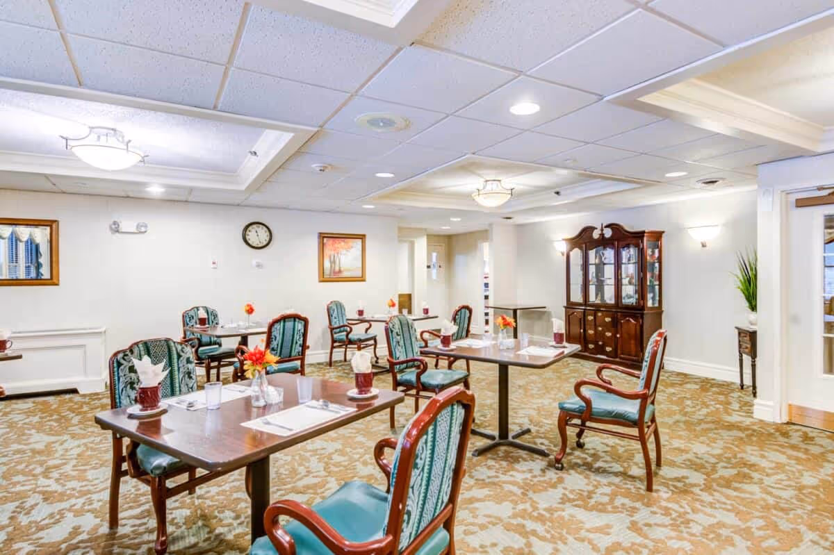 Bright dining room with several tables and upholstered chairs set for meals and a wooden china cabinet against the wall.