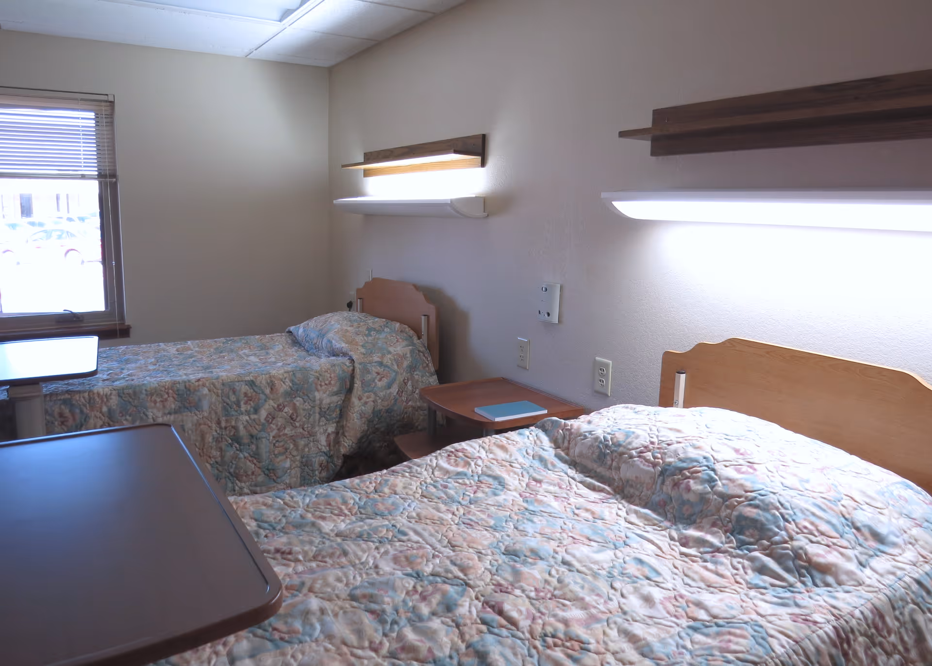 A small room with two single beds covered in floral patterned bedspreads. Each bed has a wooden headboard and a wall-mounted light fixture above it. There is a small wooden nightstand between the beds with a blue item on it. A window with blinds is visible on the left side, letting in natural light.