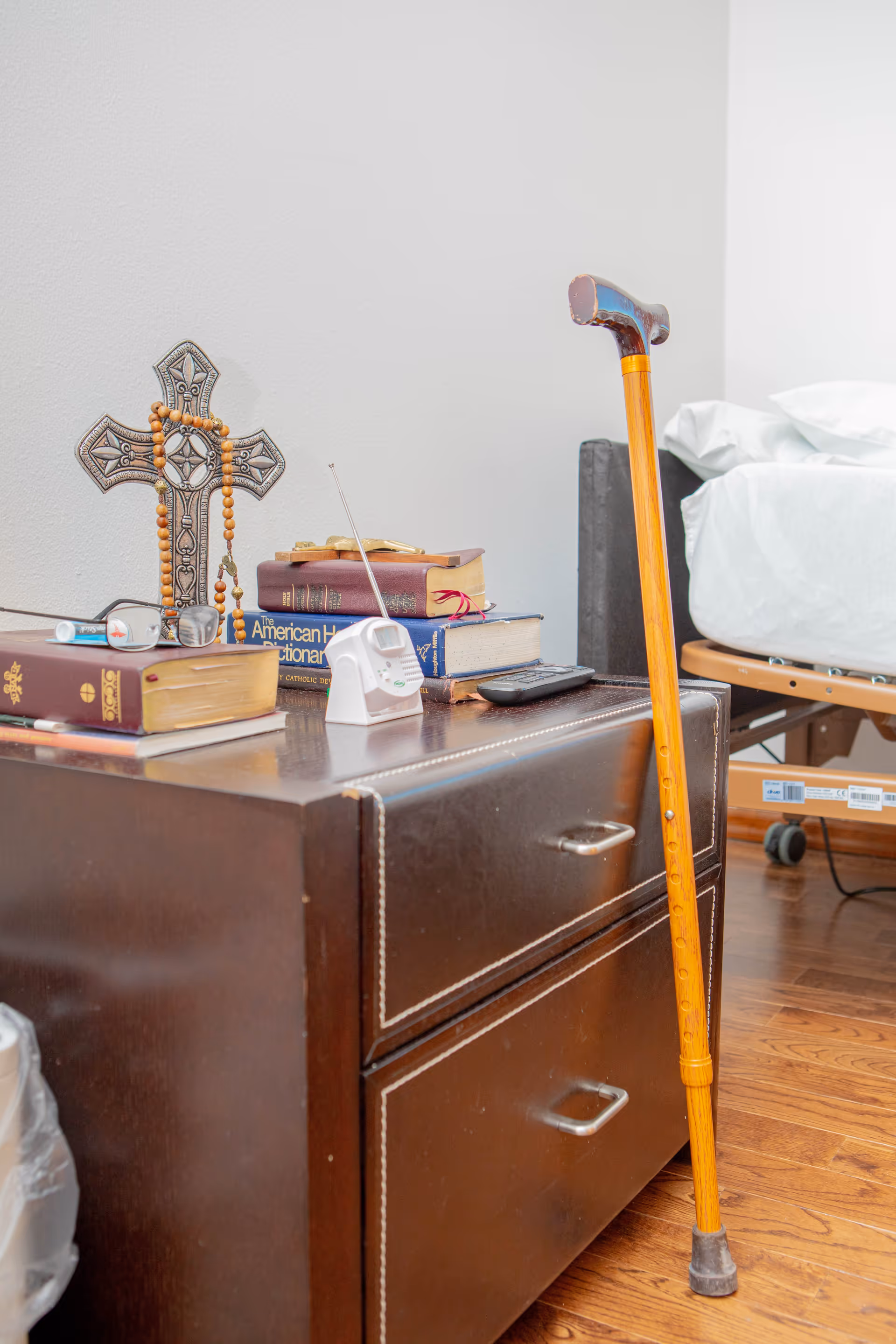 A bedside table with religious items including a decorative cross with a wooden rosary, several books, a pair of glasses, a small white radio, and a TV remote. Next to the table is a wooden cane leaning against it, and part of a bed with white bedding is visible in the background.