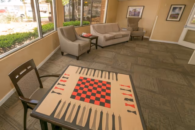 A bright lounge with a board game table in the foreground and sofas and armchairs along large windows.