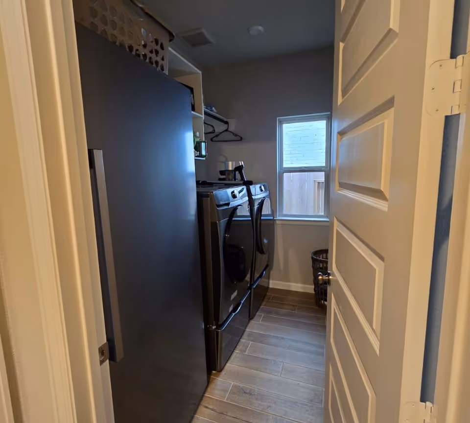 A small laundry room with a front-loading washer and dryer side by side, a window letting in natural light, a laundry basket on the floor, and shelves above the machines holding various items including a laundry basket on top of a black refrigerator.