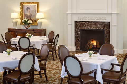 Elegant dining room with round and square tables covered in white tablecloths, each set with napkins and small floral centerpieces. The room features a lit fireplace with a marble surround, a sideboard with two lamps and a floral arrangement, and a framed portrait hanging above it. The chairs have dark wooden frames with light upholstered backs and seats. The carpet has a floral pattern.
