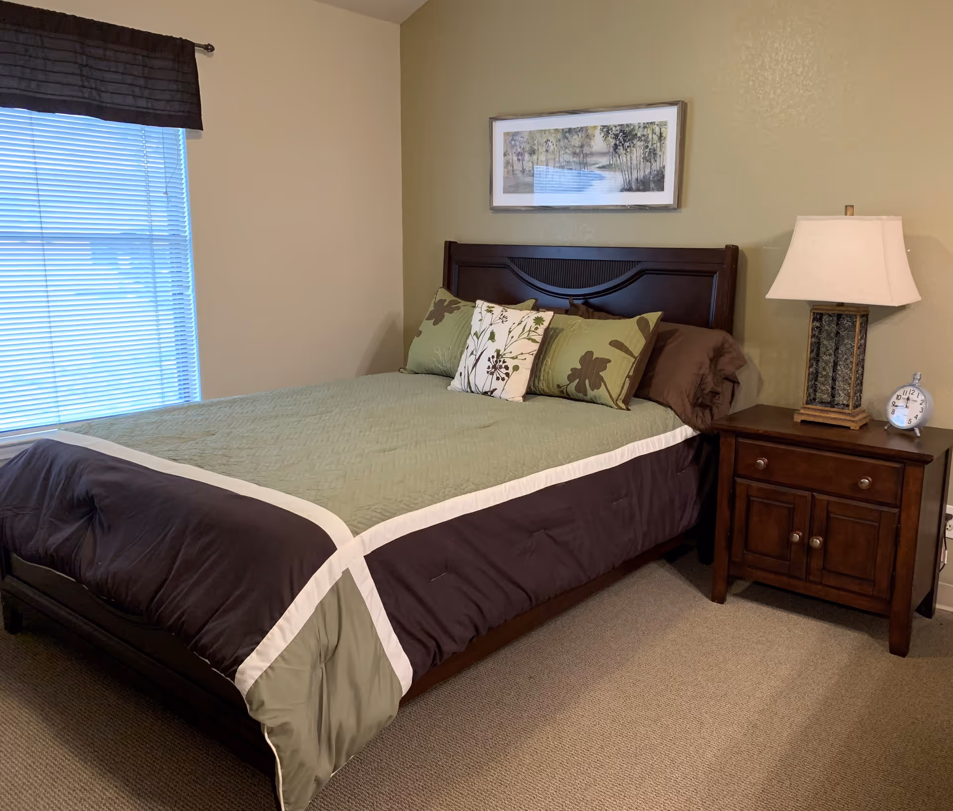 A bedroom with a neatly made bed featuring a green and brown comforter and decorative pillows. There is a wooden headboard and a nightstand with a lamp and a small clock. A window with closed blinds and a dark valance is on the left wall, and a framed landscape painting hangs above the bed.