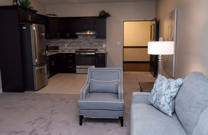 Open-concept living area with a gray sofa and armchair in the foreground and a small kitchen with dark cabinets and stainless steel appliances in the background.
