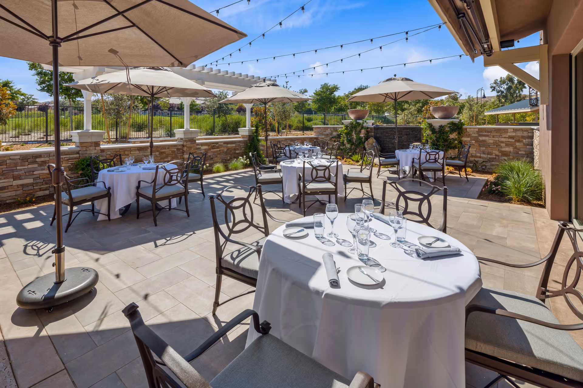 Outdoor patio dining area with round tables covered in white tablecloths, set with plates, glasses, and napkins. Several large umbrellas provide shade, and string lights are hung overhead. The area is surrounded by a low stone wall and greenery under a clear blue sky.