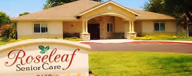 Front exterior view of Roseleaf Senior Care facility, a single-story building with a covered entrance, stone pillars, and a well-maintained lawn with a sign displaying the facility's name.
