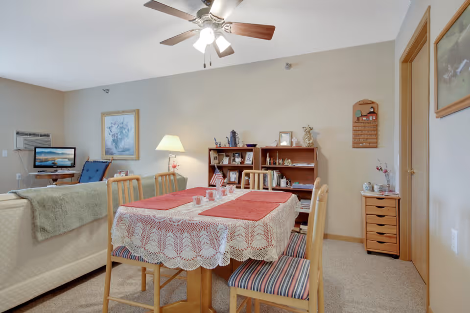 A cozy living and dining area in a senior living facility with a wooden dining table covered with a lace tablecloth and red placemats, surrounded by four wooden chairs with striped cushions. Behind the table is a wooden bookshelf with various decorative items and framed photos. To the left, there is a beige couch with a green throw blanket, a rocking chair, a lamp, and a small TV on a stand. The walls are light beige with framed artwork, and there is a ceiling fan with lights above.