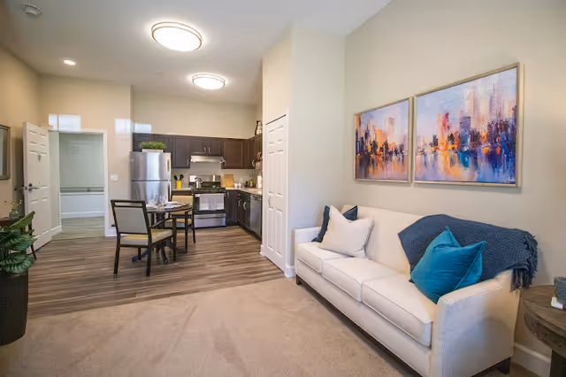 Interior view of a senior living facility apartment featuring a small kitchen with stainless steel appliances, a dining table with two chairs, and a living area with a white sofa adorned with blue and white pillows and a blue throw blanket. Two colorful abstract paintings hang on the wall above the sofa.