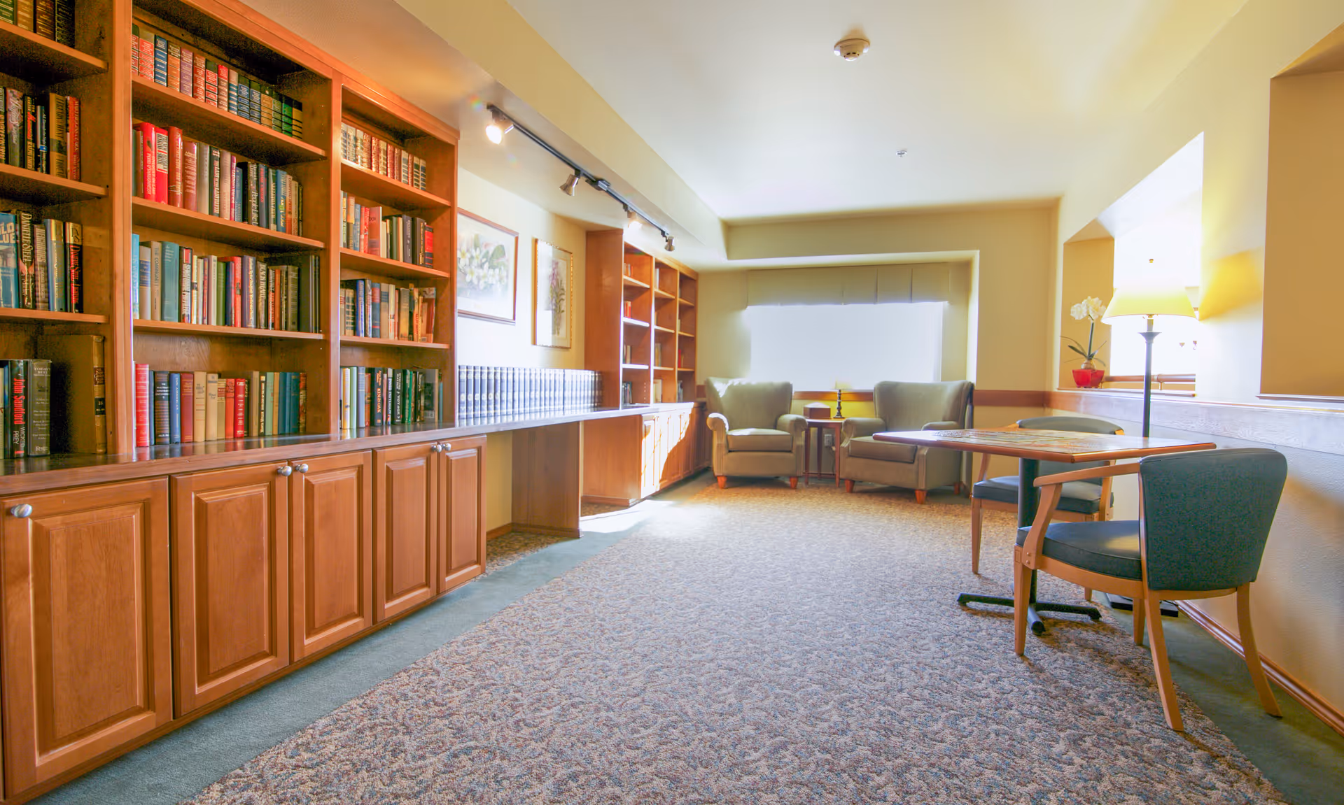 A cozy reading and sitting area in a senior living facility with wooden bookshelves filled with books along the left wall, two armchairs near a window with bright natural light, a small table between the chairs, and a table with two chairs and a lamp on the right side.