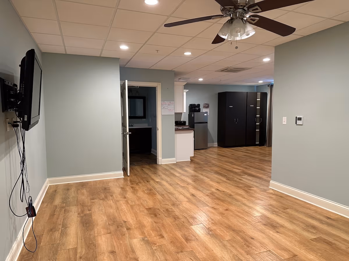 Empty senior living common area with wood-look flooring, a ceiling fan, wall-mounted TV, small kitchenette and dark storage cabinets.