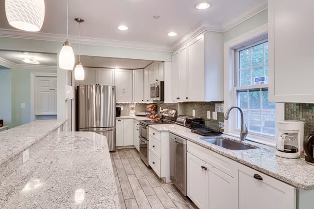 Bright and modern kitchen with white cabinets, granite countertops, stainless steel refrigerator, oven, microwave, dishwasher, and a window above the sink. Pendant lights hang over a breakfast bar with seating.