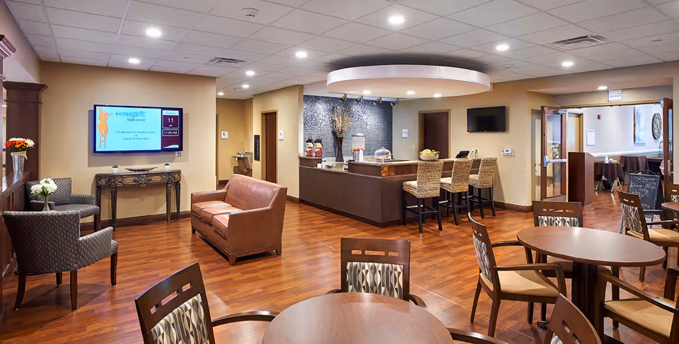 A spacious senior living common area with wooden floors, round tables with chairs, a leather couch, and two armchairs. There is a wall-mounted TV displaying a welcome message, a counter with bar stools, and a decorative wall with vases and a lamp. The room is well-lit with recessed ceiling lights and has an open doorway leading to another room.