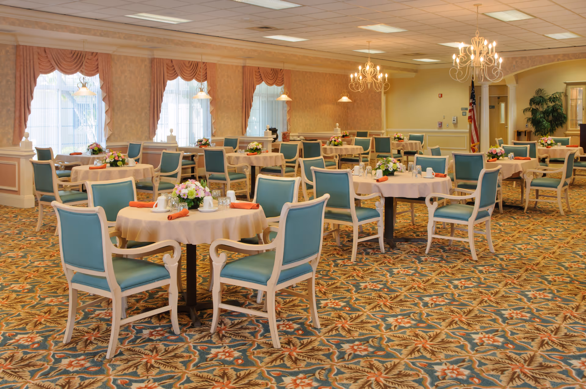A spacious dining room with multiple round tables covered with beige tablecloths, each set with cups, glasses, and orange napkins. The chairs have light wood frames and teal upholstery. The room features large windows with peach-colored drapes, chandeliers hanging from the ceiling, and a patterned carpet with floral designs.