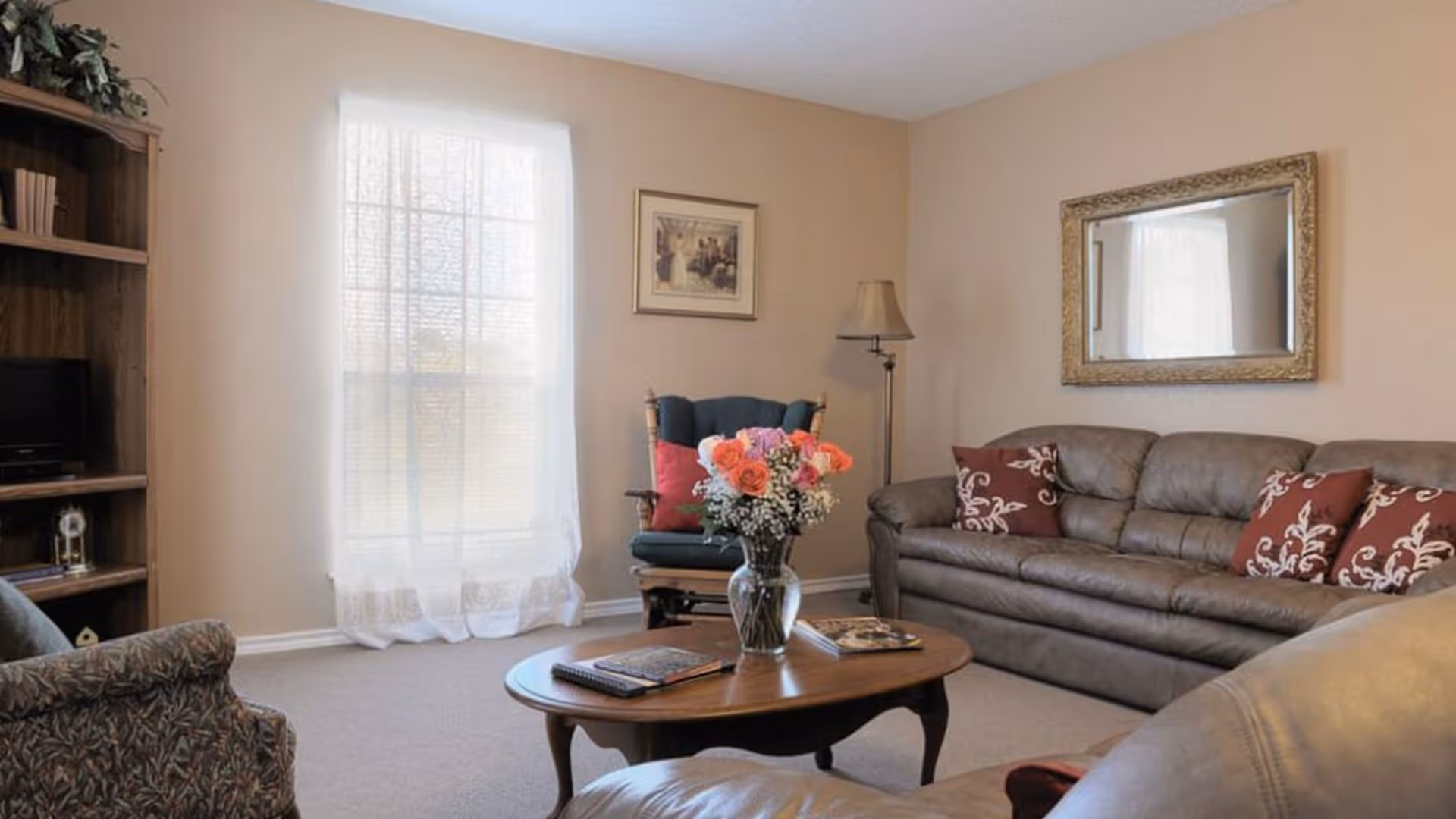 A cozy living room with leather sofas and armchairs arranged around a wooden coffee table topped with a vase of flowers, a floor lamp, and a bookcase by a sheer curtained window.