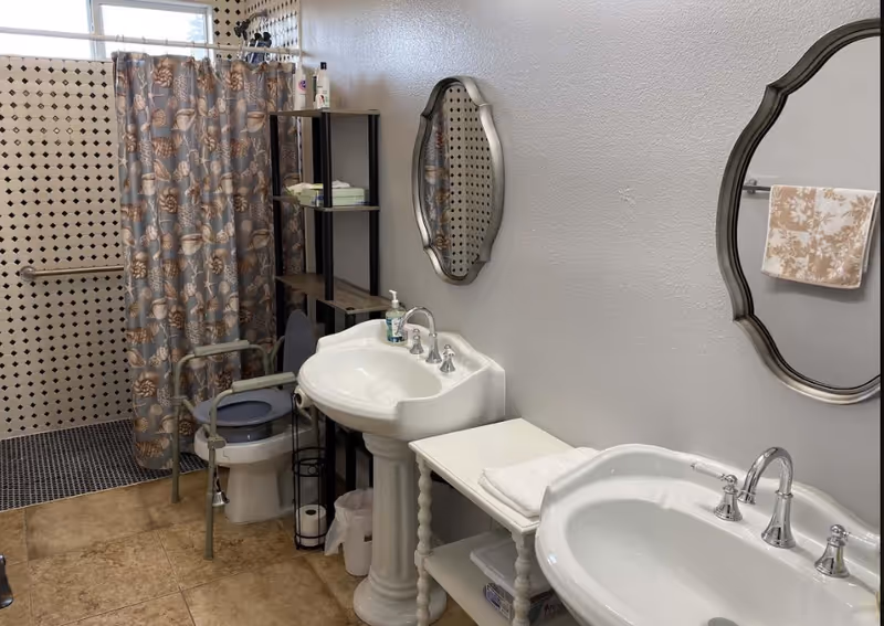 A bathroom with two white pedestal sinks, each with a decorative mirror above. There is a toilet with a safety frame around it and a shower area with a floral-patterned shower curtain. A black shelving unit holds toiletries and other items. The walls have a white and black tile pattern near the shower, and the floor is tiled in a beige color. A towel hangs on a rack on the wall.