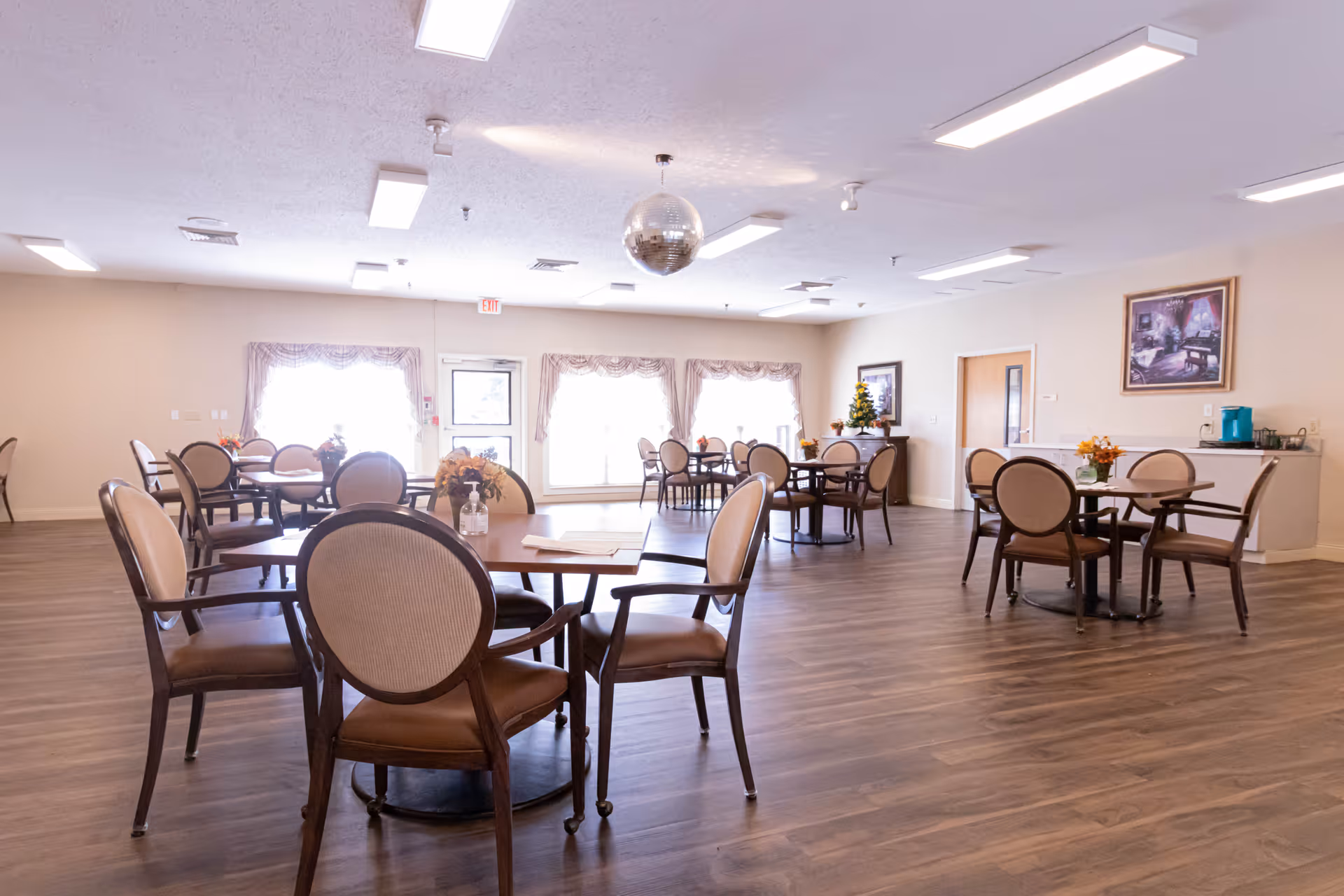 Spacious senior living dining room with round tables and chairs on wood flooring and large windows.