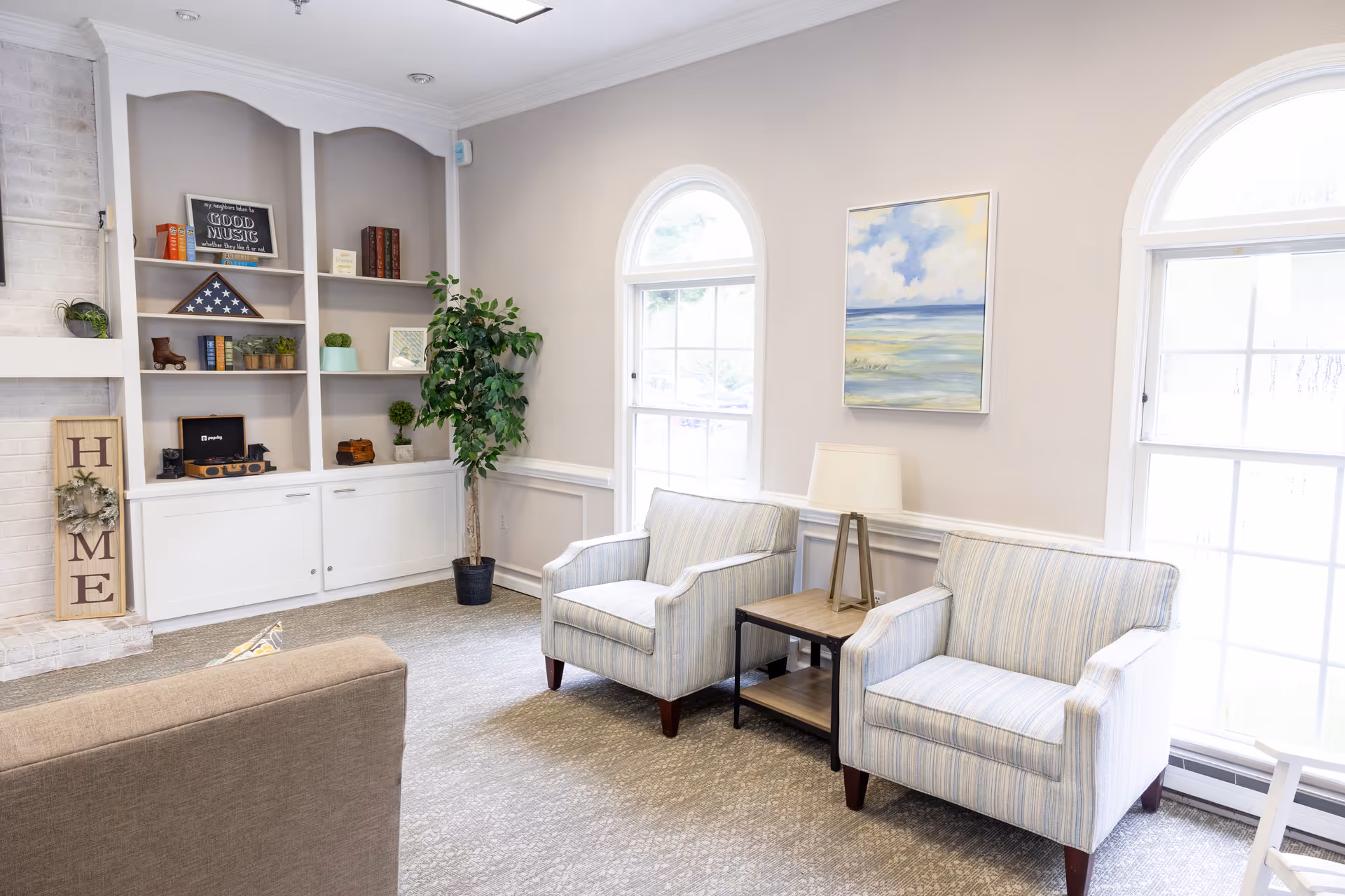 A bright and cozy living room area with two striped armchairs separated by a wooden side table with a lamp. There are two large arched windows letting in natural light. A built-in white shelving unit holds books, decorative items, and a folded American flag. A tall potted plant is placed next to the shelving. The walls are painted light beige, and a painting of a seascape hangs above the armchairs.