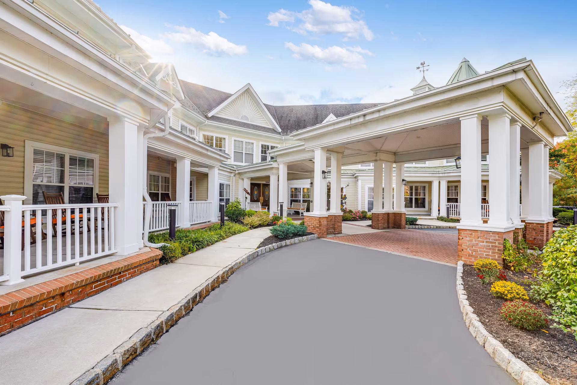 Covered porte-cochere and front entrance of a light-colored senior living building with white columns, a porch, and landscaped walkway.
