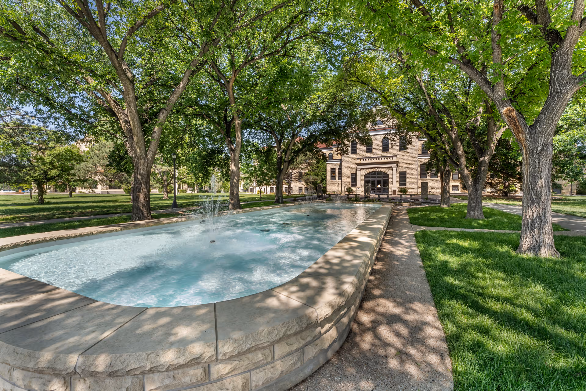 A long rectangular water fountain surrounded by a stone border is situated in a park-like area with green grass and large leafy trees. In the background, there is a stone building with arched windows and a central entrance. The scene is bright and sunny with shadows from the trees cast on the ground and water.