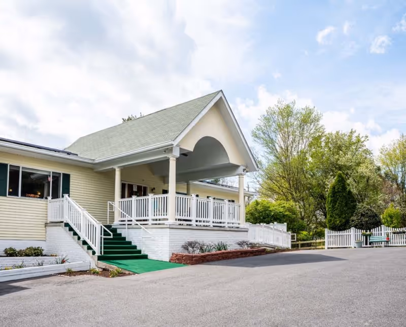 Exterior view of a single-story building with a covered porch, white railings, and green carpeted stairs leading up to the entrance. The building is surrounded by trees and a paved driveway.